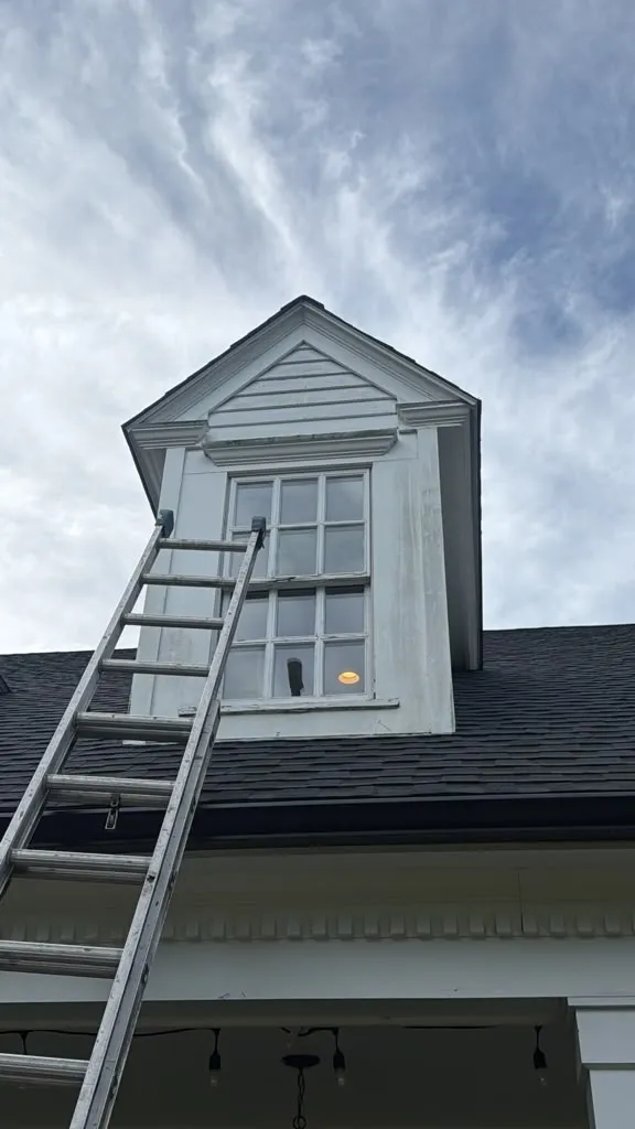 Original white dormer window with divided panes and weathered exterior trim, with a ladder leaning against the dormer.dormer window replacement in Hanover, MA