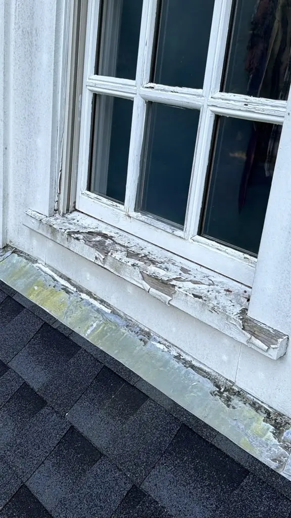 Close-up of an old dormer window with peeling white paint and deteriorated wood along the sill and surrounding trim.