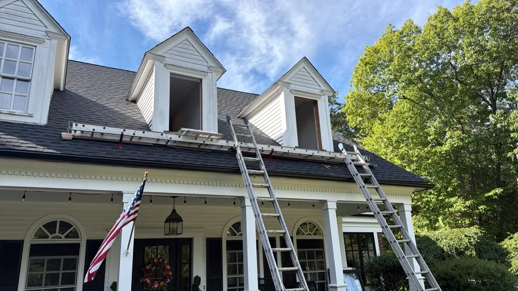 Two open dormer window openings above a front porch, with ladders positioned for window replacement.