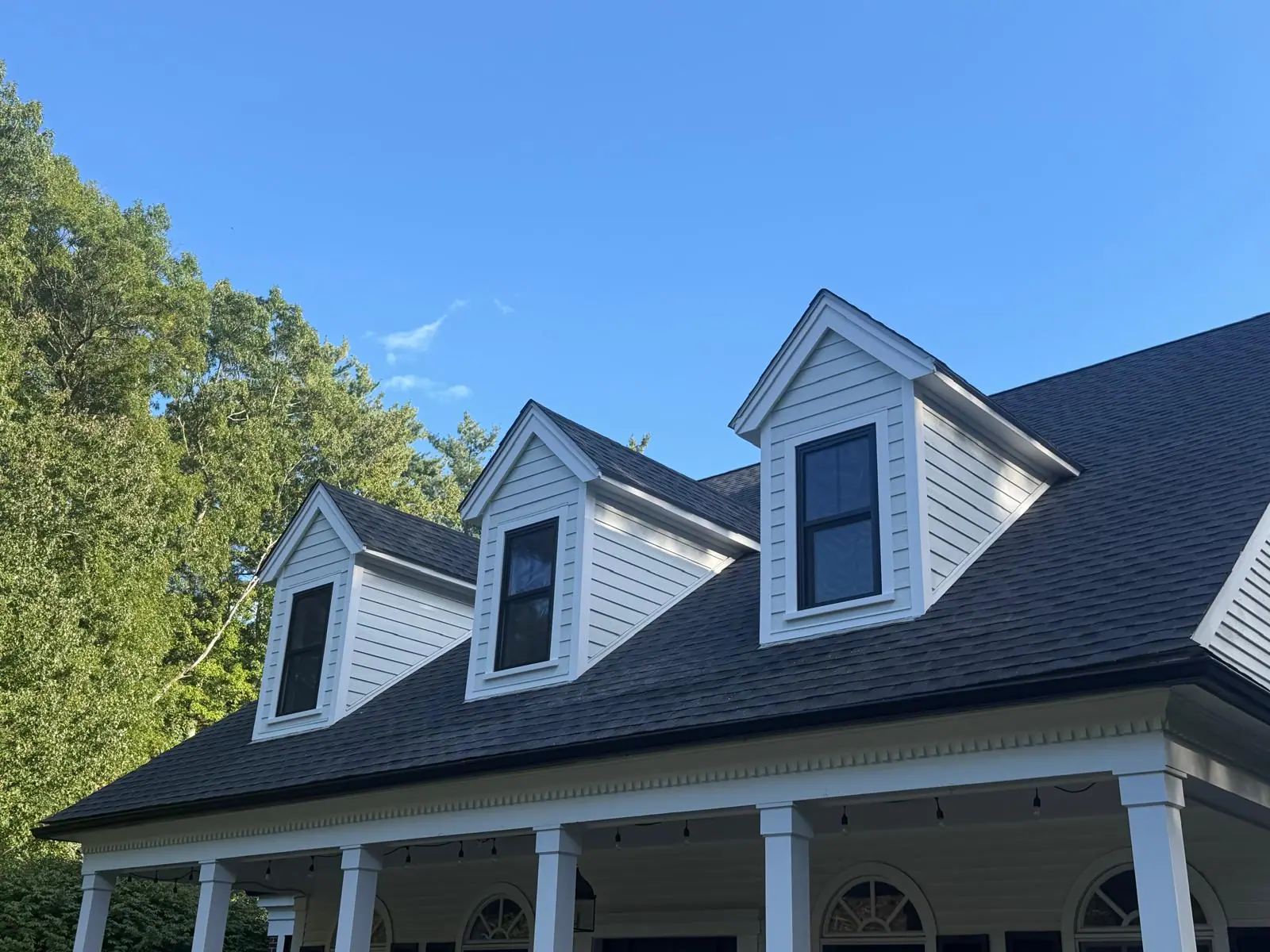 Angled view of three finished dormers with black window frames, white trim, and white siding above a shingled roof.